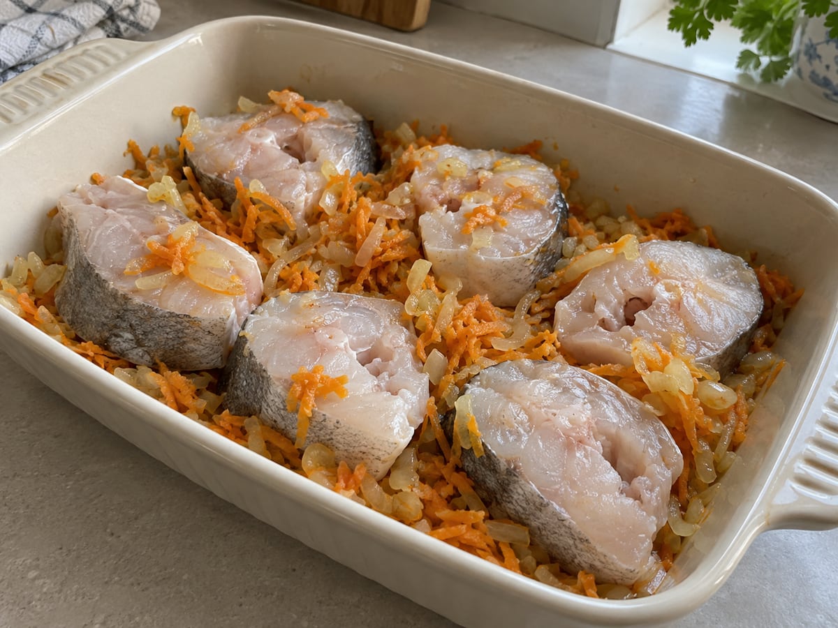 Сырые куски хека уложены на слой лука и моркови в форме